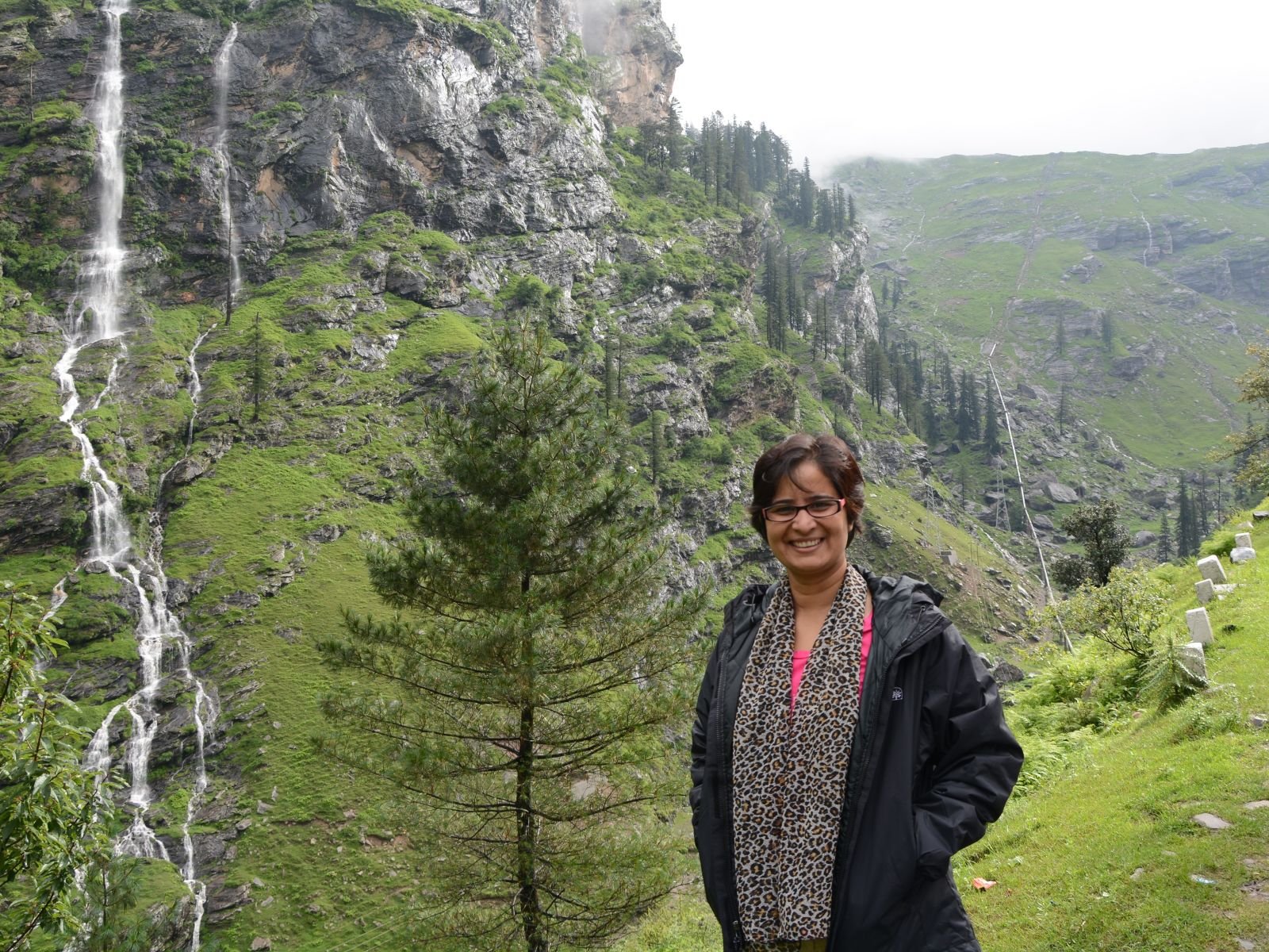 Manu standing beside a green mountain road and waterfall in Lahaul, Himachal Pradesh